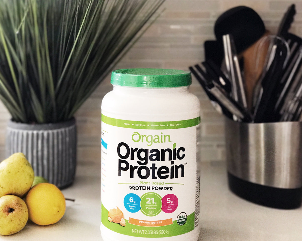 A container of Orgain Organic Protein powder, plant-based in peanut butter flavor, is placed on a kitchen counter next to a bunch of yellow pears. In the background, a plant, a knife block, and various kitchen utensils are visible.