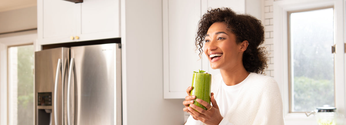 A smiling woman holding a green smoothie in a bright and modern kitchen. She has curly hair and is dressed in a white top. Behind her, there is a stainless steel refrigerator and a large window letting in natural light.