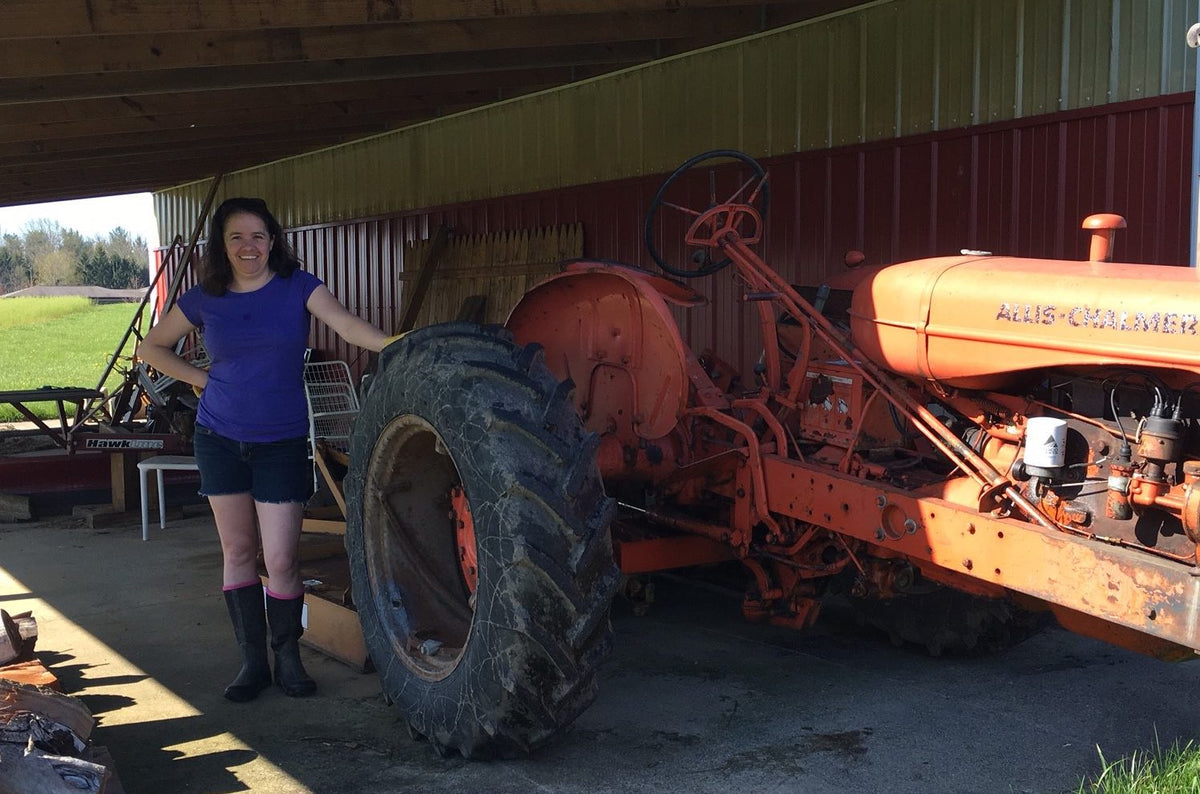 A woman wearing a purple shirt, black shorts, and black boots stands next to an old orange tractor under a shed. She has one hand on the large rear tire of the tractor and is smiling at the camera. There is a farm landscape in the background.