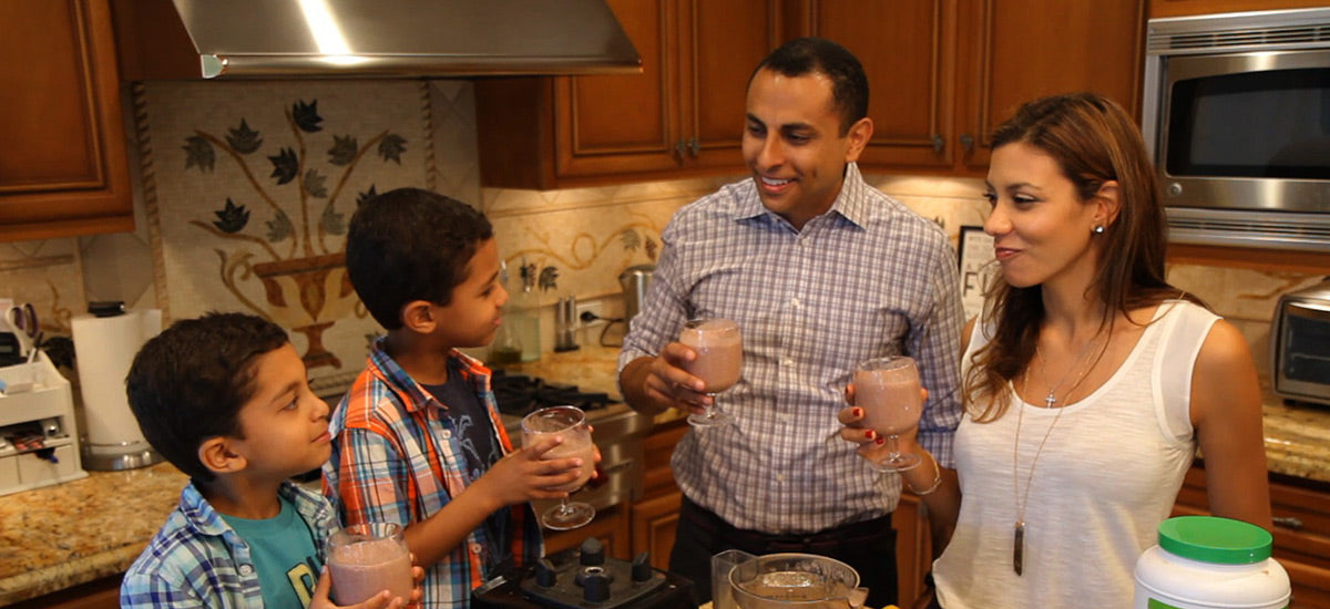 A family of four smiles and enjoys smoothies in a kitchen with wooden cabinets, a blender, and various kitchen items.