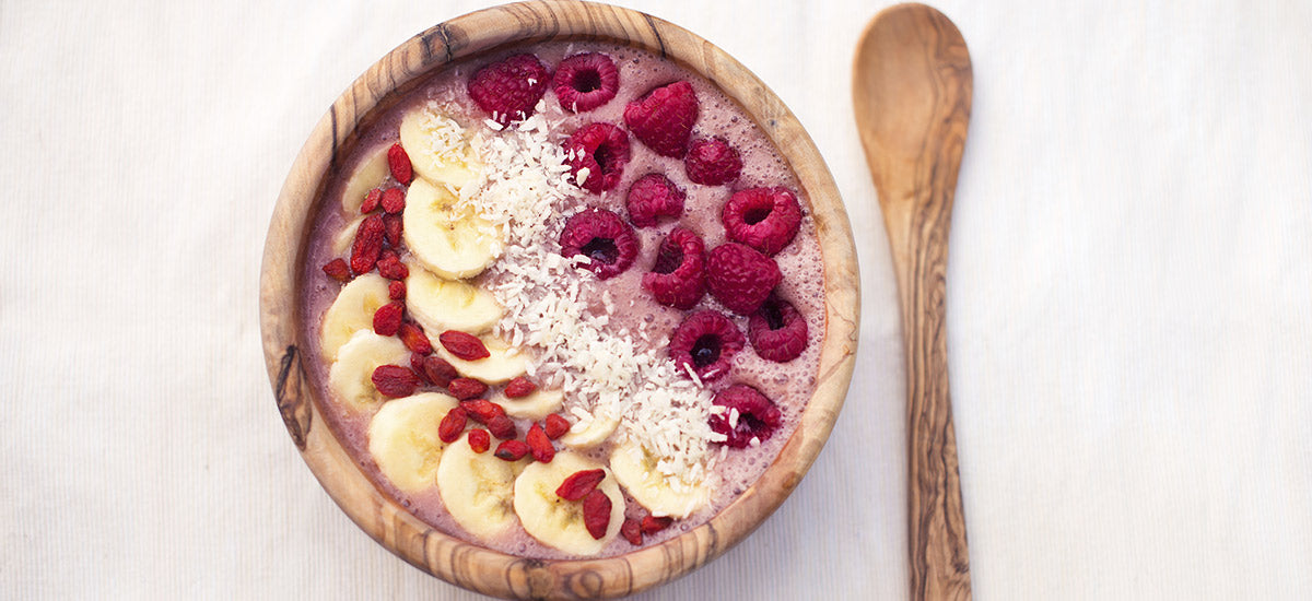 A wooden bowl of smoothie topped with sliced bananas, raspberries, goji berries, and shredded coconut, placed on a light-colored cloth next to a wooden spoon. The smoothie is pink, and the toppings are neatly arranged in rows.