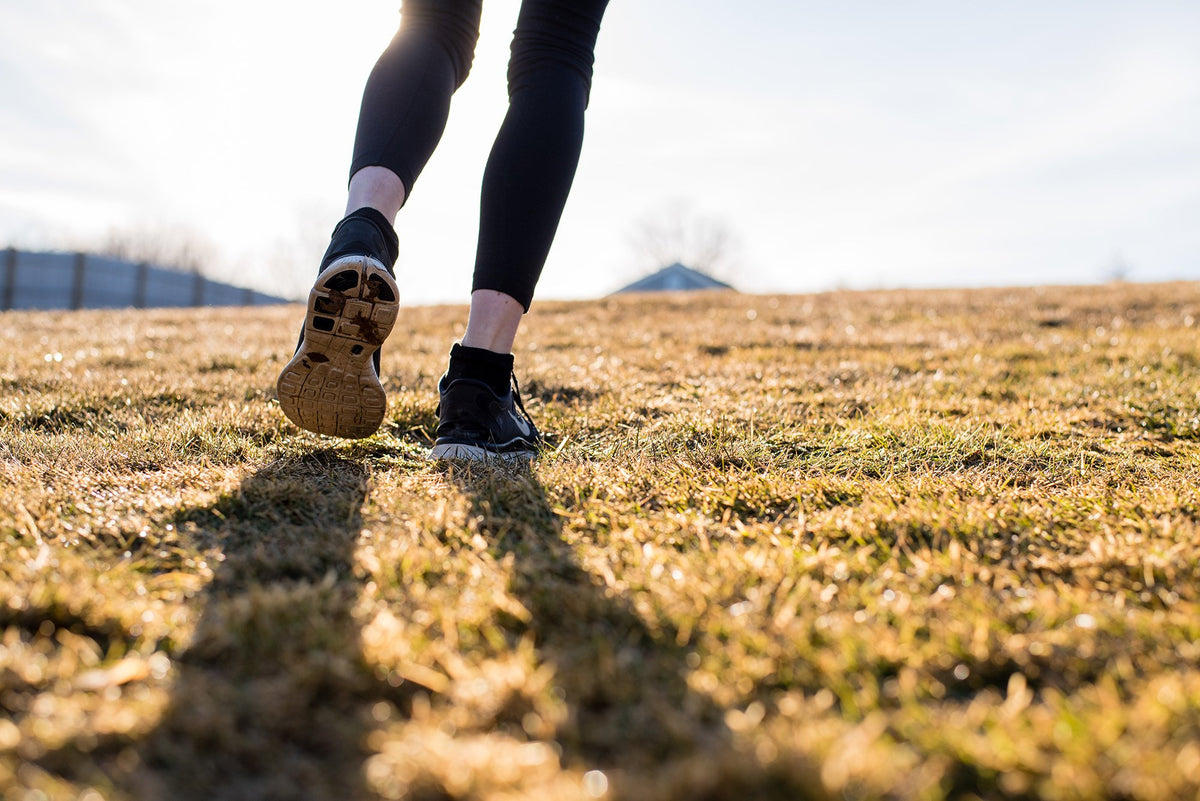 Close-up of a person in black leggings and running shoes jogging or walking on dry grass with the sun casting long shadows in the early morning or late afternoon. The background is slightly blurred, featuring a building and trees in the distance.