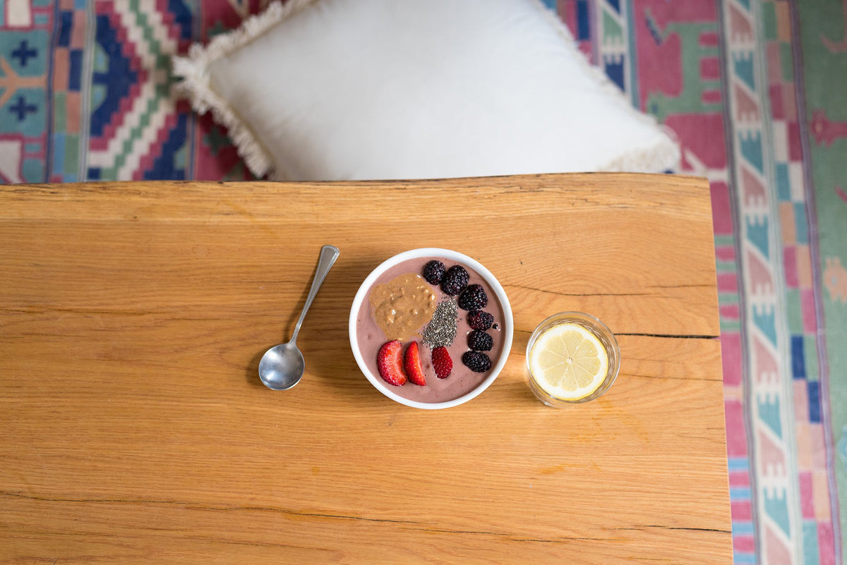 A wooden table holds a bowl of smoothie topped with strawberries, blackberries, chia seeds, and nut butter. Beside the bowl is a spoon and a glass of water with a lemon slice. A colorful rug and a white cushion are partially visible in the background.