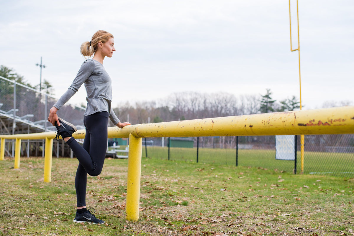 A woman is stretching her quadriceps by pulling her right foot toward her buttock while balancing her left hand on a yellow metal fence at an outdoor field. She is wearing a gray long-sleeve top, black leggings, and black sneakers.