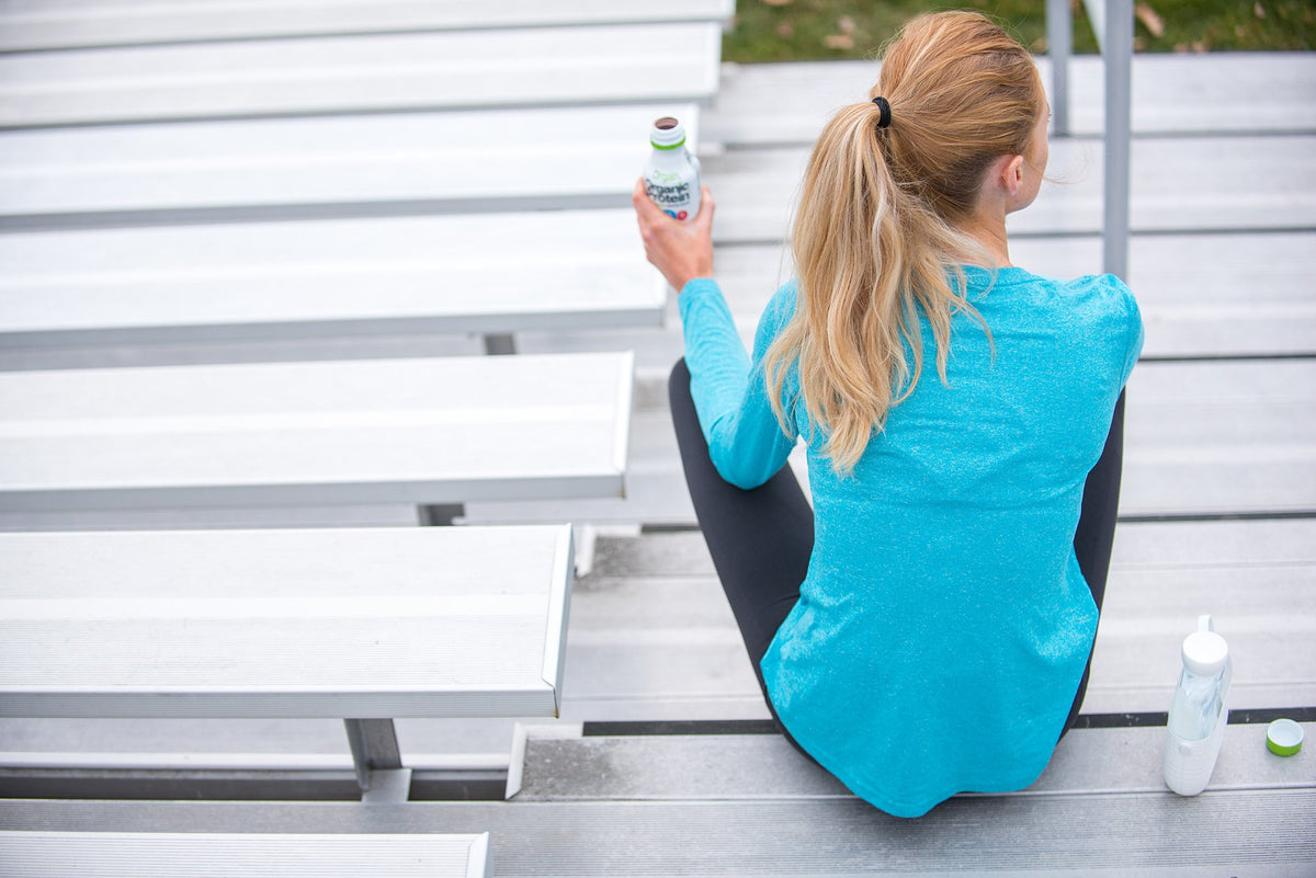 A person with blonde hair, tied in a ponytail, wearing a blue long-sleeve shirt and black leggings, is sitting on gray bleachers. They are holding a bottle in one hand and have two other bottles placed nearby. The setting appears to be outdoors.