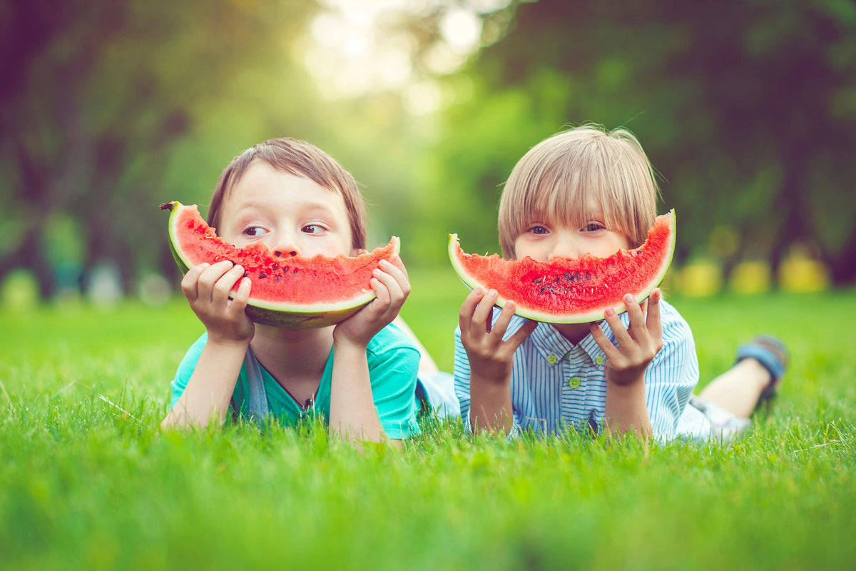 Two children lying on the grass hold large watermelon slices in front of their faces, smiling.