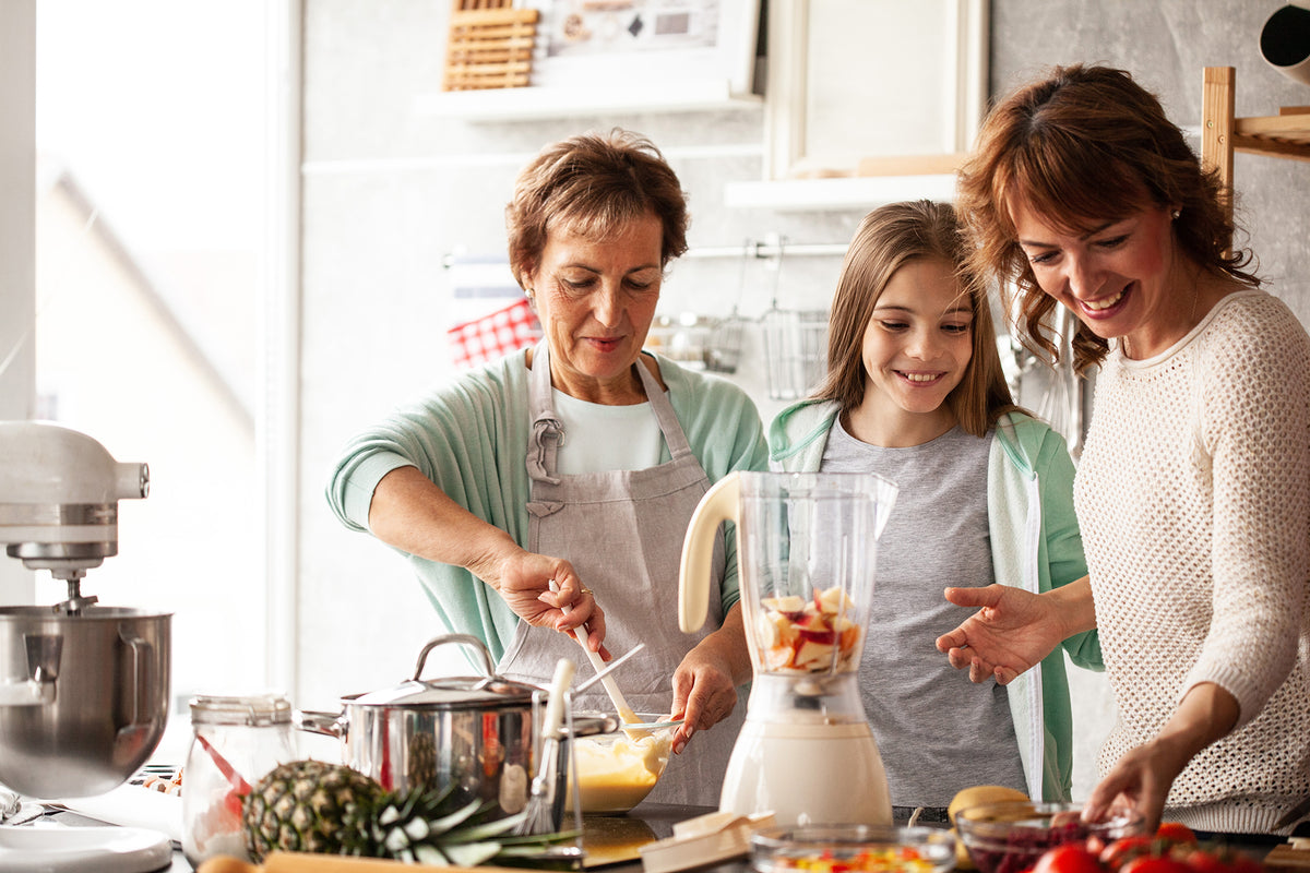 Three people in a bright kitchen, cooking together. An older person stirs ingredients, a child smiles, and an adult cuts fruit.