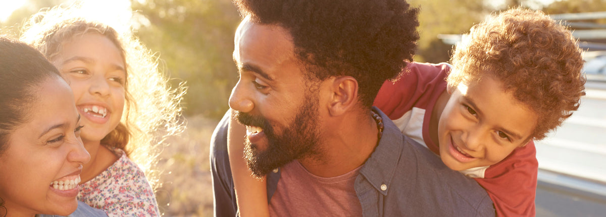 A happy family enjoys an outdoor moment. An adult man carries a smiling child on his back, while an adult woman and another child, both smiling and cheerful, are close by. The sun shines brightly in the background, creating a warm, glowing atmosphere.