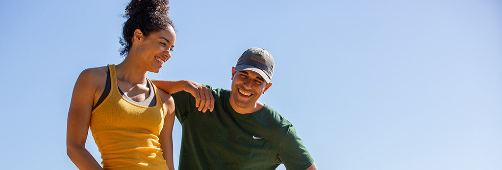 A woman in a yellow tank top and a man in a green shirt with a baseball cap are outdoors, smiling and looking at each other against a clear blue sky. They appear to be enjoying a warm, sunny day.