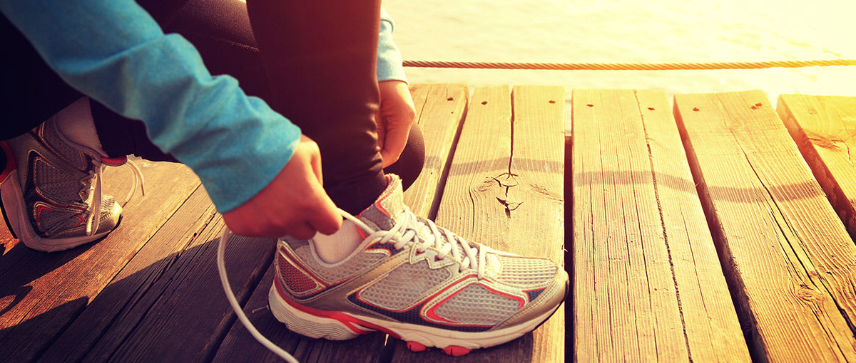 A person wearing a blue long-sleeve shirt ties the laces of a grey and pink running shoe while crouched on a wooden boardwalk. Sunlight casts a warm glow on the scene.
