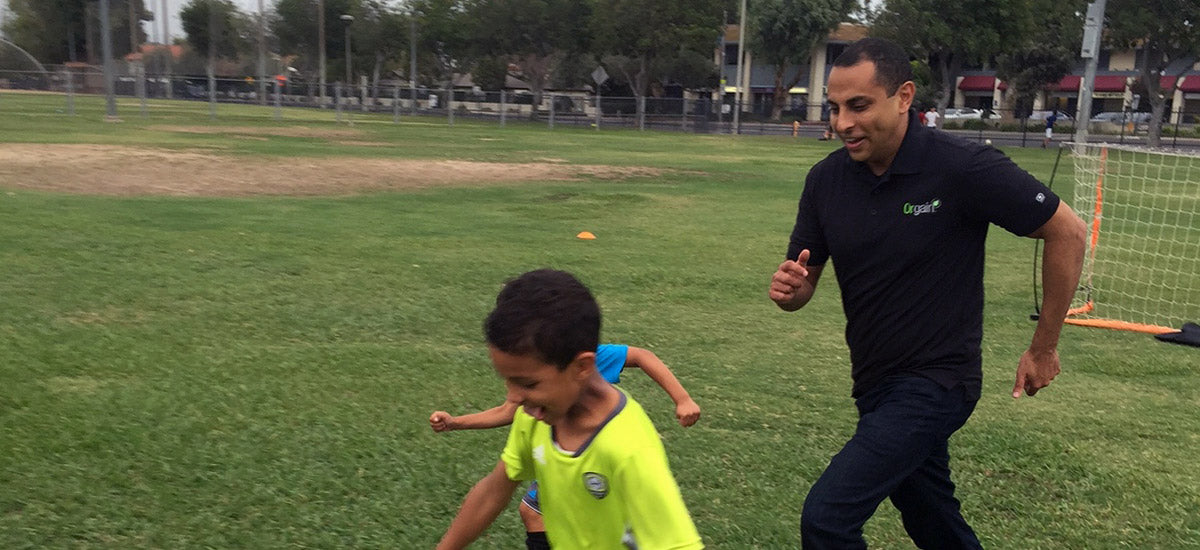 A man and two children are playing soccer on a grassy field. The children are running ahead of the man, who is also running and smiling. A small soccer goal is visible in the background, along with trees and a few buildings. The day appears to be cloudy.