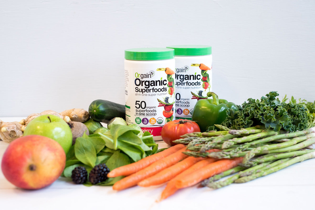 Two containers of Orgain Organic Superfoods powder are surrounded by assorted fresh produce, including carrots, asparagus, green bell peppers, kale, zucchini, apples, peaches, spinach, ginger, and blackberries. The background is a light-colored surface.