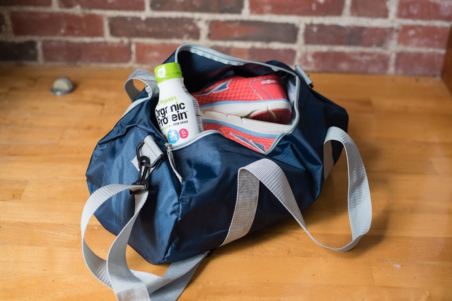 A blue gym bag with gray straps is open on a wooden floor, revealing a pair of red and white athletic shoes and a bottle of organic protein drink. The protein bottle is partially tucked inside the bag. In the background, there is a brick wall.