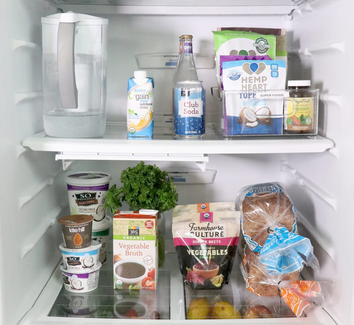 An open refrigerator contains a water filter pitcher, club soda, hemp seeds, a jar of pickles, containers of probiotic yogurt, vegetable broth, mixed veggies, parsley, a loaf of whole grain bread, lemons, and a basket with garlic cloves.