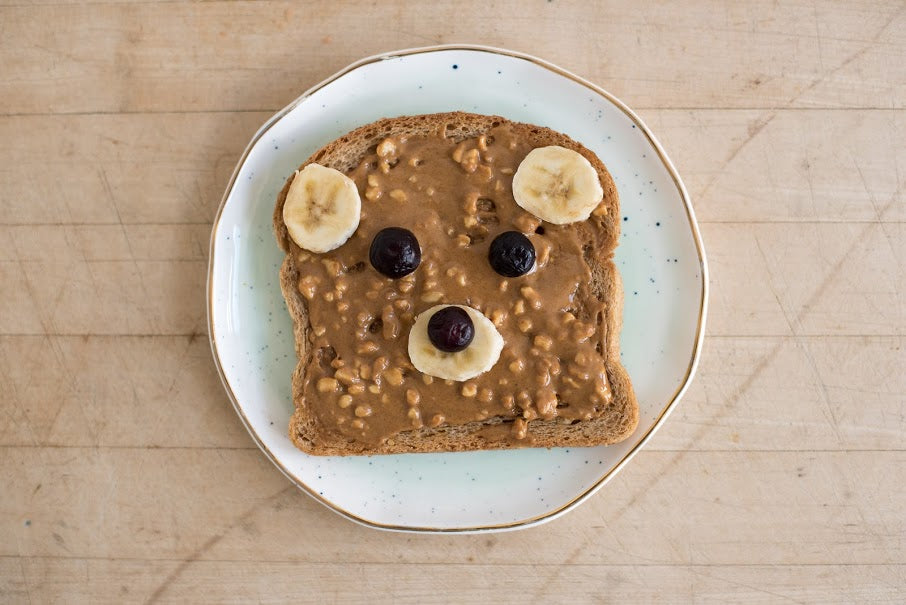 A slice of toast with peanut butter, banana slices, and blueberries arranged to look like a bear's face on a white plate.