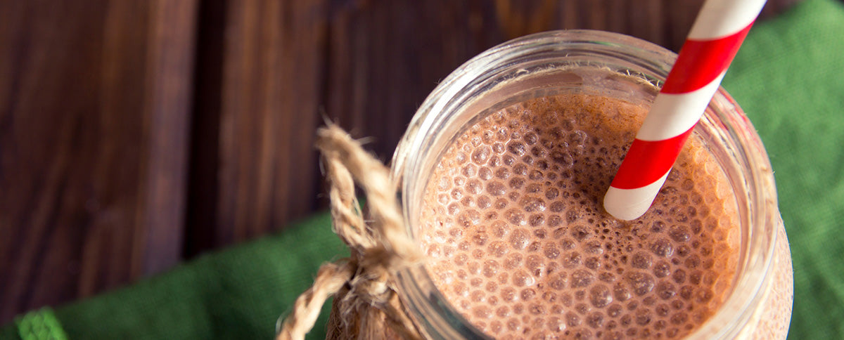 A close-up of a bubbling chocolate milkshake in a glass jar topped with a red and white striped straw. The jar is tied with a rustic string bow, and a green cloth is partially visible underneath, placed on a wooden surface.