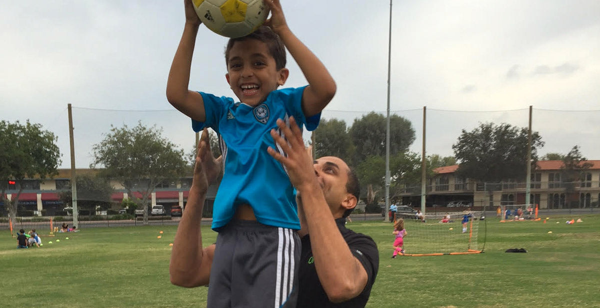 A man holds up a smiling boy who is lifting a soccer ball above his head. They are on a grassy field with other people and soccer equipment visible in the background under a cloudy sky.