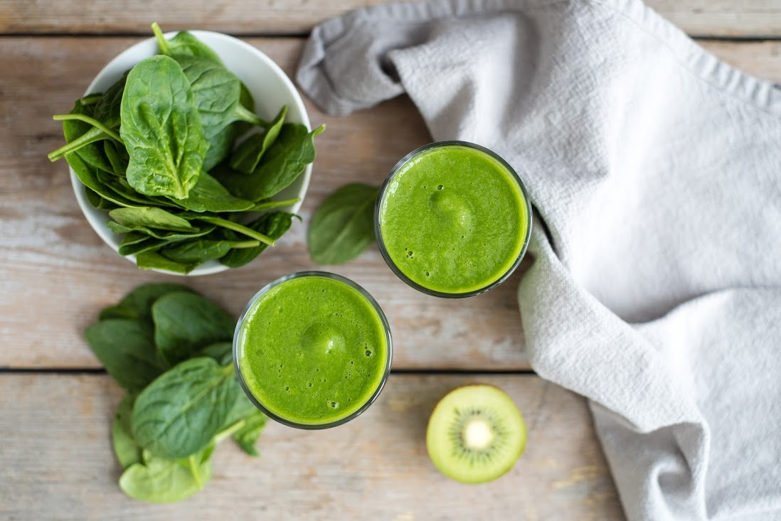 A top-down view of two glasses filled with green smoothies on a wooden table. A bowl of fresh spinach leaves, a halved kiwi, and a gray cloth are placed nearby, along with spinach leaves scattered on the table.