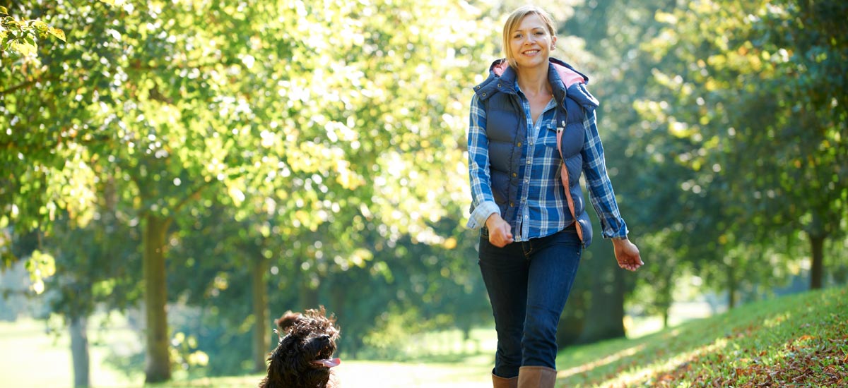 A woman in a blue and white plaid shirt and a navy vest is walking a curly-haired brown dog along a sunlit, tree-lined path in a park. She looks happy and relaxed, enjoying the stroll on a clear day. The background includes lush green trees and grass.