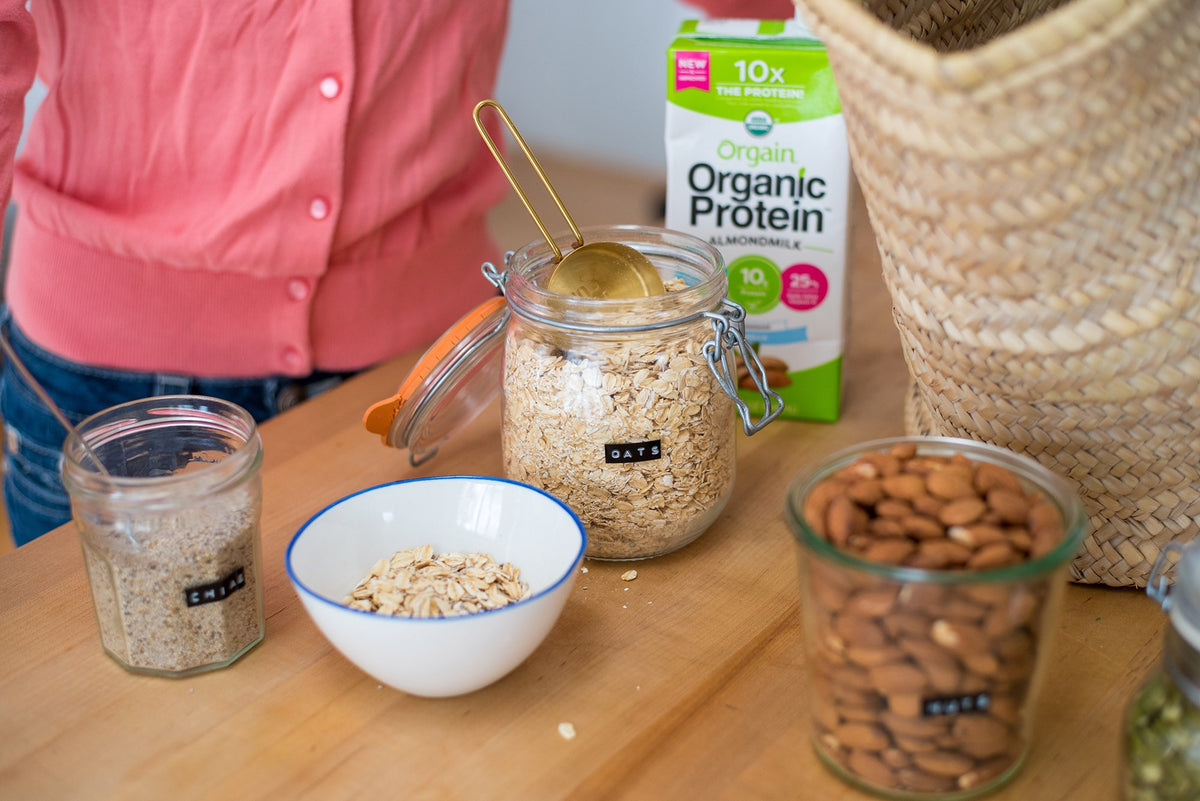 A person in a pink cardigan is standing next to a table with various food items, including a jar labeled "OATS," a jar labeled "CHIA," a bowl of oats, a jar of almonds, a gold-colored scoop, a carton of organic almond milk protein, and a woven basket.