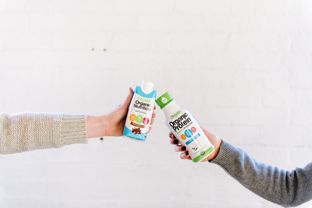 Two hands are pictured holding different Orgain nutritional beverages against a white brick wall. The left hand holds a carton of Orgain Organic Nutrition in chocolate flavor, and the right hand holds a bottle of Orgain Organic Protein in vanilla flavor.