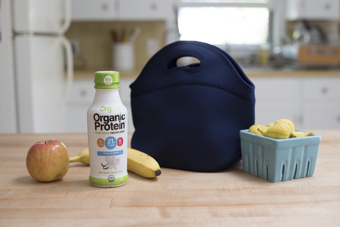 An Orgain Organic Protein shake next to a lunchbag and fresh fruit.