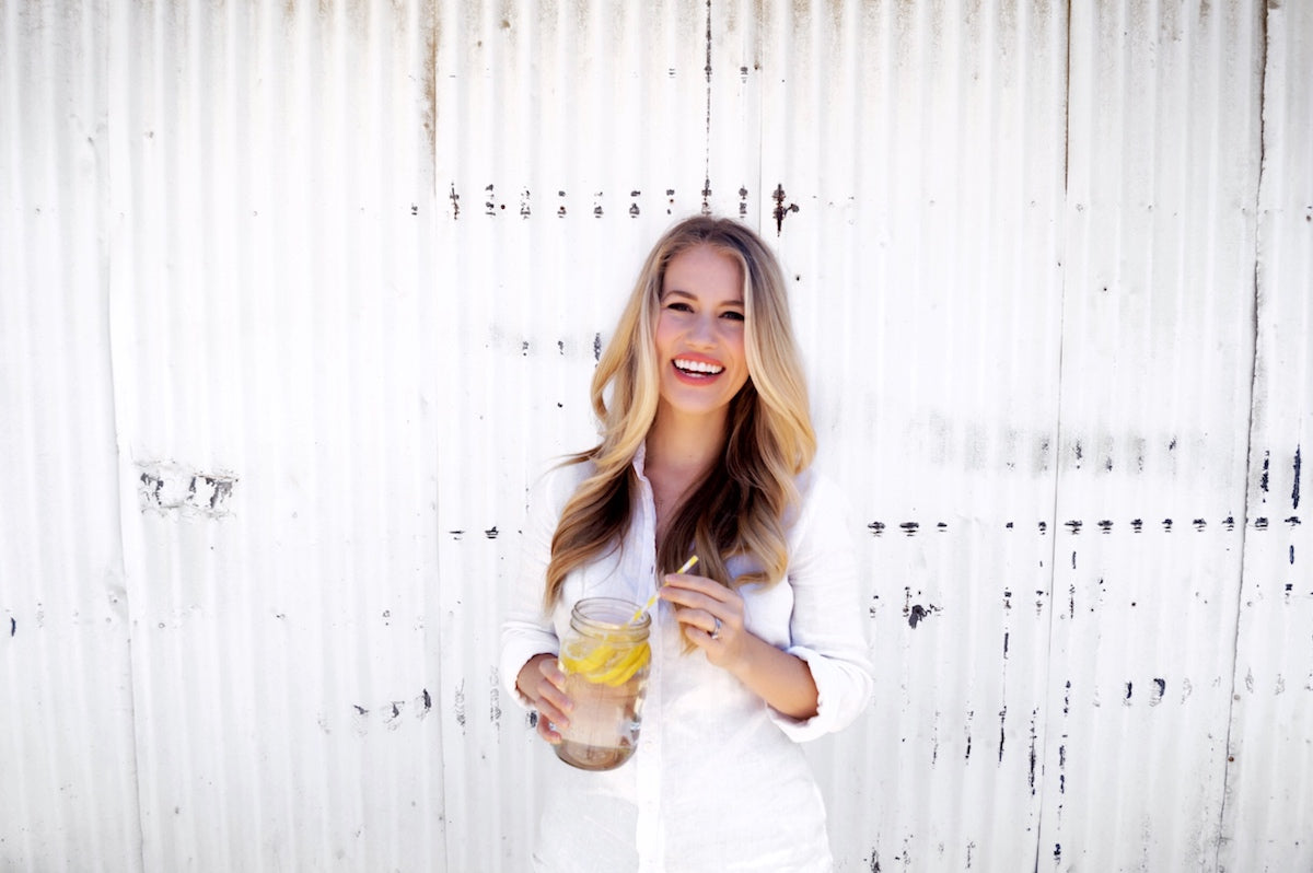 A person with long blonde hair smiles while holding a mason jar filled with a lemon beverage. They are standing in front of a white corrugated metal wall and wearing a white shirt.