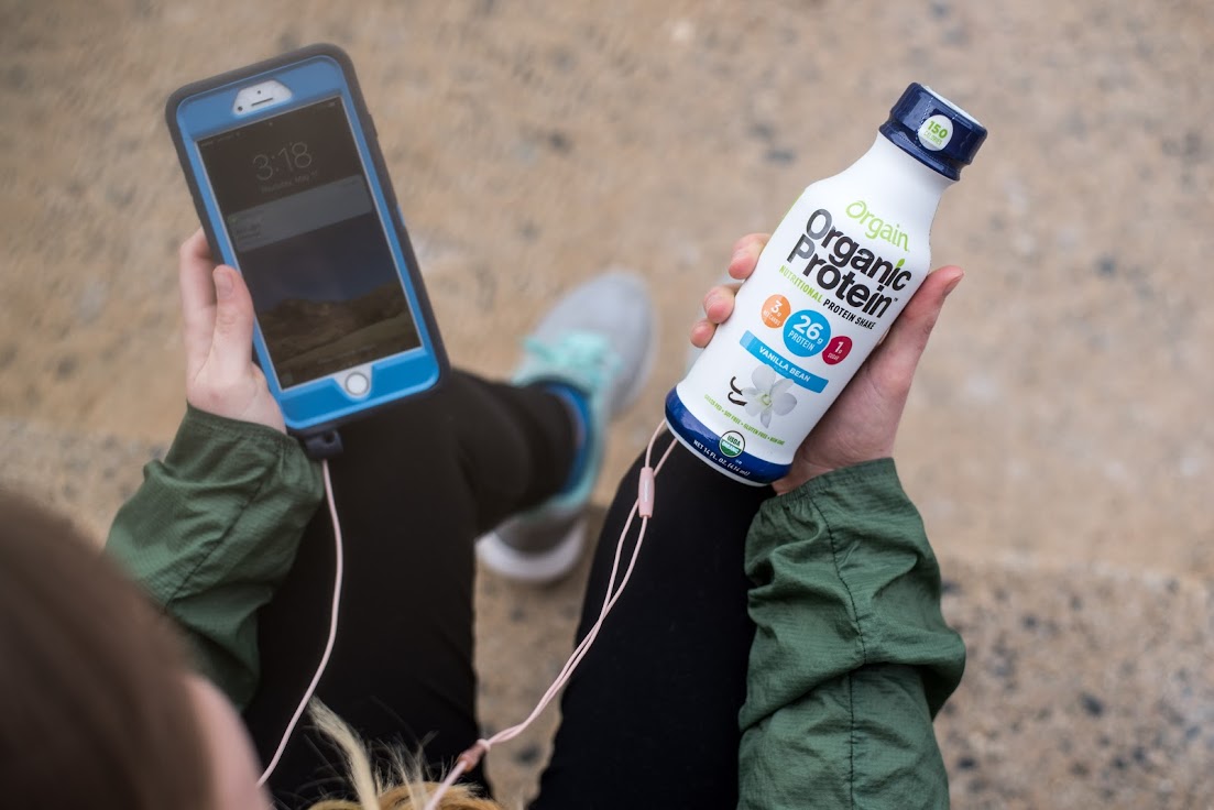 A person wearing a green jacket and black leggings holds a bottle of Orgain Organic Protein shake in one hand and a smartphone in the other. The phone has blue and grey casing with earbuds attached. The background shows a sandy surface.