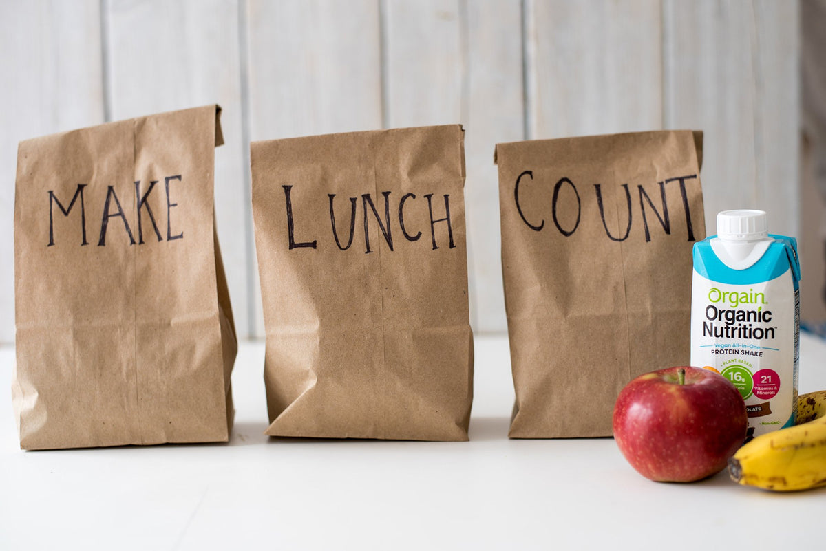 Three brown paper bags with ‚ÄúMAKE‚Äù on the first, ‚ÄúLUNCH‚Äù on the second, and ‚ÄúCOUNT‚Äù on the third. To the right, there is a carton of Orgain Organic Nutrition protein shake, an apple, and bananas. The background is a light wooden wall.