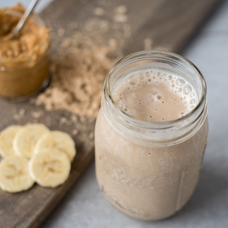 A mason jar filled with a creamy, frothy beige smoothie is placed on a wooden board. Behind the jar, there is a small bowl of peanut butter with a spoon and several banana slices arranged on the board. Powder is sprinkled around the bowl.