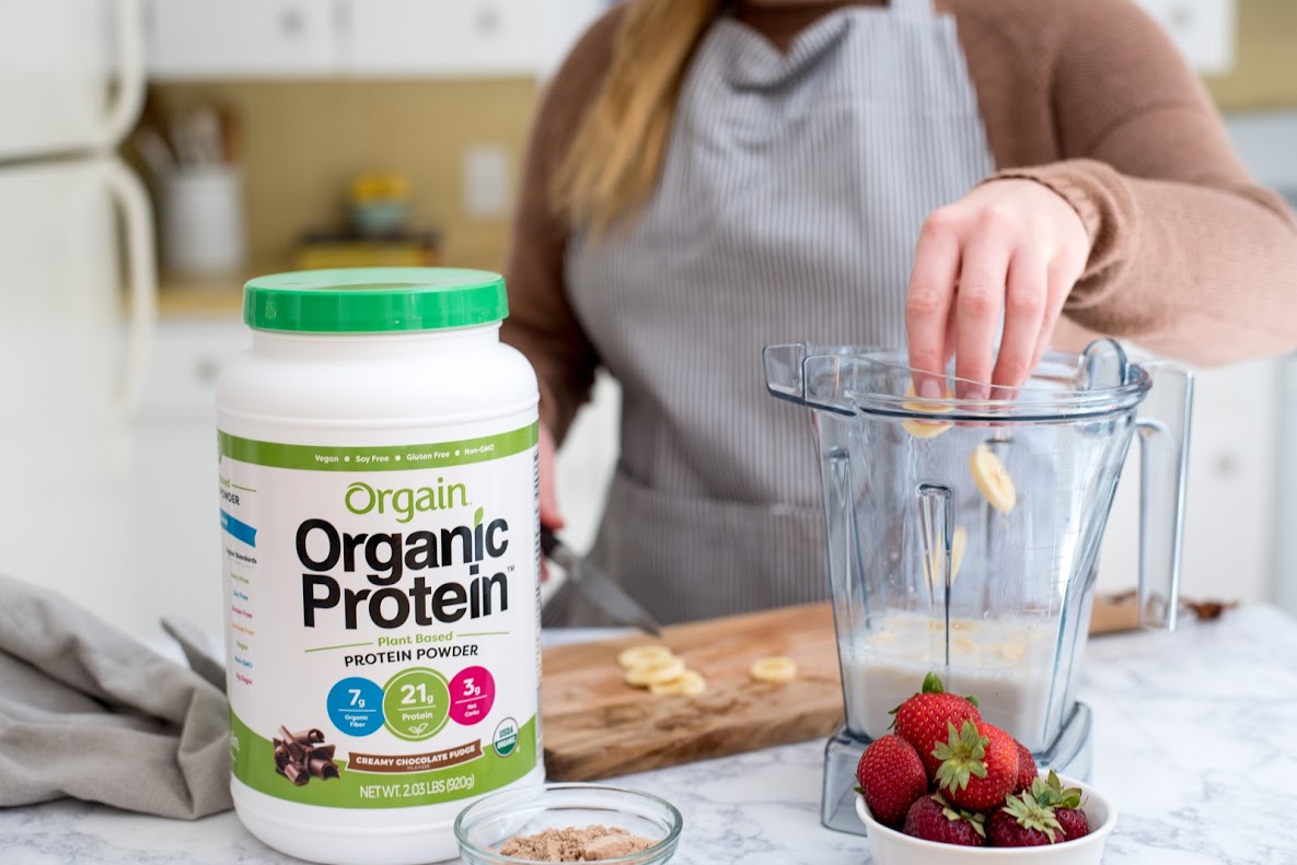 A person wearing a striped apron is preparing a smoothie. They are adding banana slices into a blender. A container of Orgain Organic Protein powder is on the counter beside strawberries, a towel, and a small bowl of protein powder.