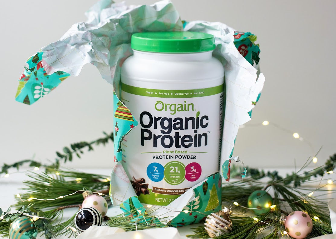 Orgain Organic Protein powder with festive wrapping among holiday decorations, highlighting 7g fiber, 21g protein, 5g net carbs.