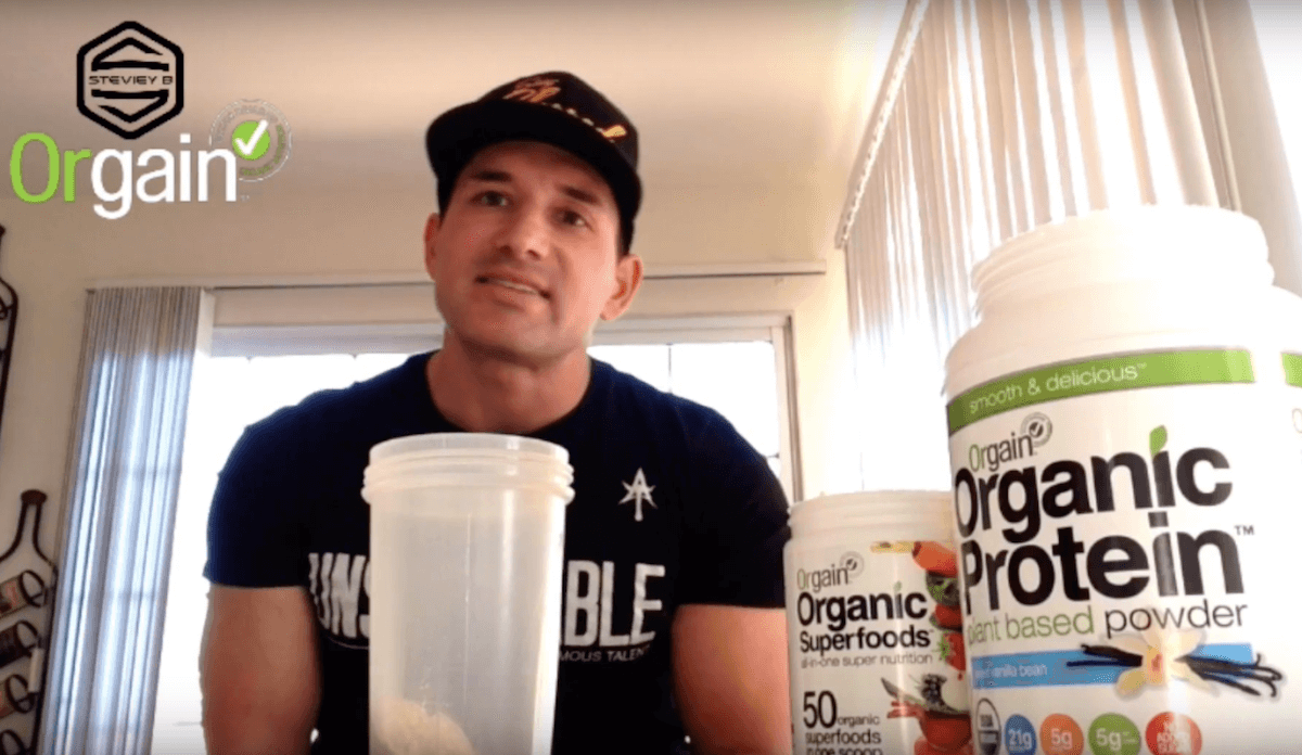 A man in a black cap is standing at a kitchen counter, preparing a shake. In front of him are containers of Orgain Organic Superfoods and Orgain Organic Protein powder. The Orgain logo and checkmark are visible in the top left corner.