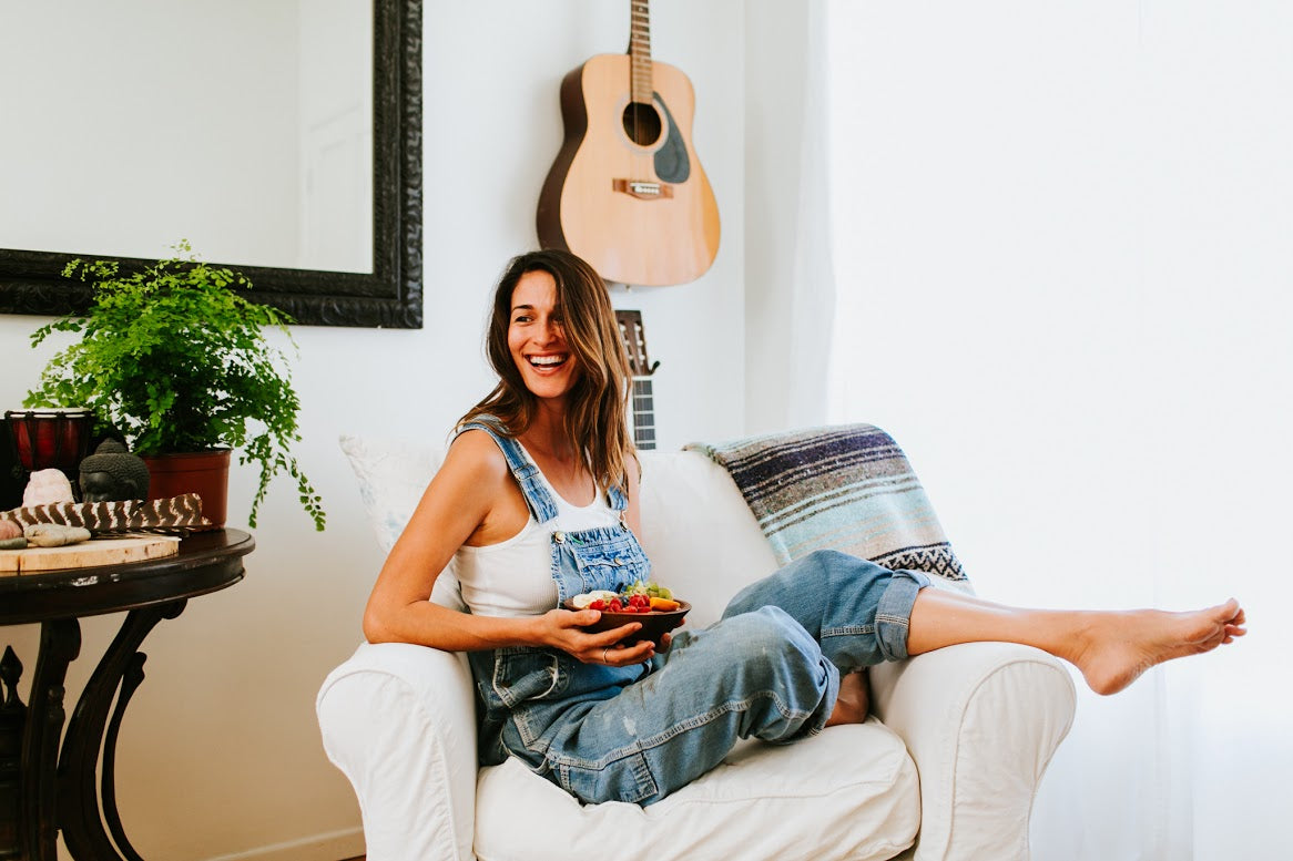 A happy woman sitting in a chair with a bowl of vegetables and fruits in her lap.