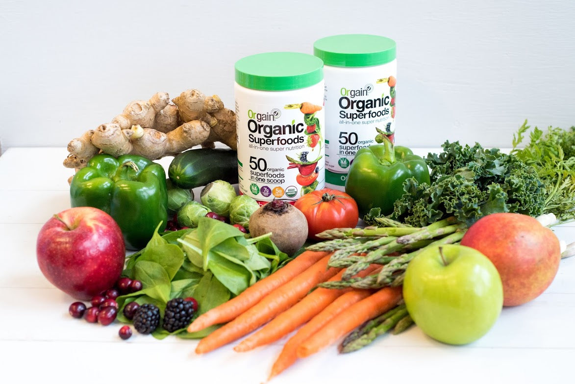 A selection of fresh fruits and vegetables, including carrots, apples, peppers, ginger, tomatoes, asparagus, spinach, berries, and kale, surrounds two containers of Orgain Organic Superfoods powder with 50 superfoods listed on the label.