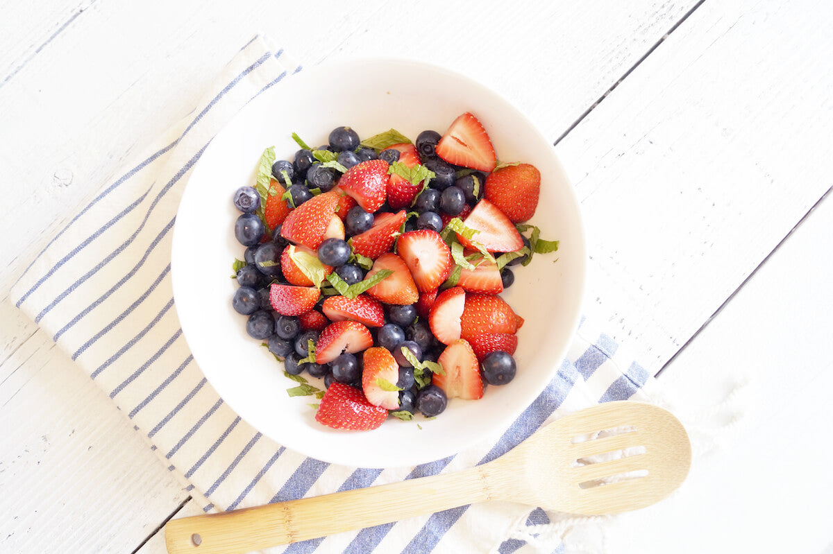 A white bowl filled with a fresh fruit salad containing blueberries, sliced strawberries, and mint leaves. The bowl is placed on a blue and white striped cloth next to a wooden spoon, all set on a white wooden surface.