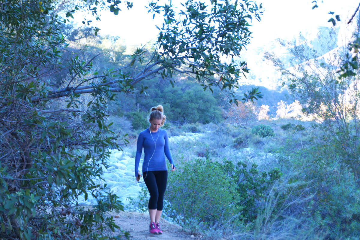 A woman wearing a blue long-sleeve shirt, black leggings, and pink shoes is walking on a trail surrounded by greenery. The background features a mix of trees, shrubs, and patches of sunlight filtering through, giving a serene and natural setting.