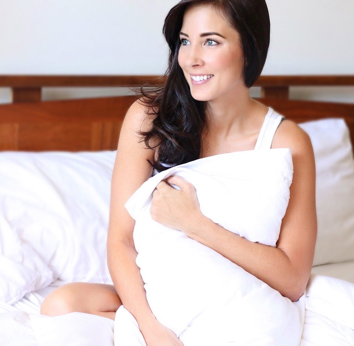 A woman with long, dark hair sits in bed wrapped in a white blanket, smiling and looking off to the side. She is on a bed with white sheets and a wooden headboard. The overall scene is bright and cozy.