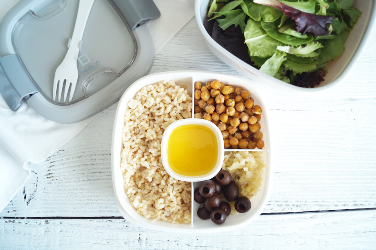 A bento box featuring compartments with brown rice, roasted chickpeas, black olives, and grated cheese, with a central container holding a yellow sauce. Next to it is a container with mixed green salad. The background is a white wooden table.