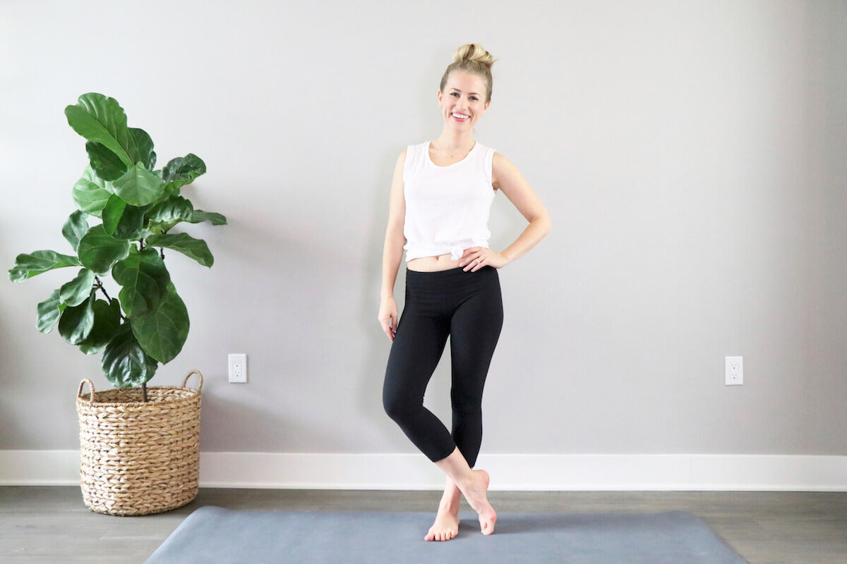 A woman stands on a blue yoga mat, posing with one hand on her hip, in a bright room with a large green plant nearby.