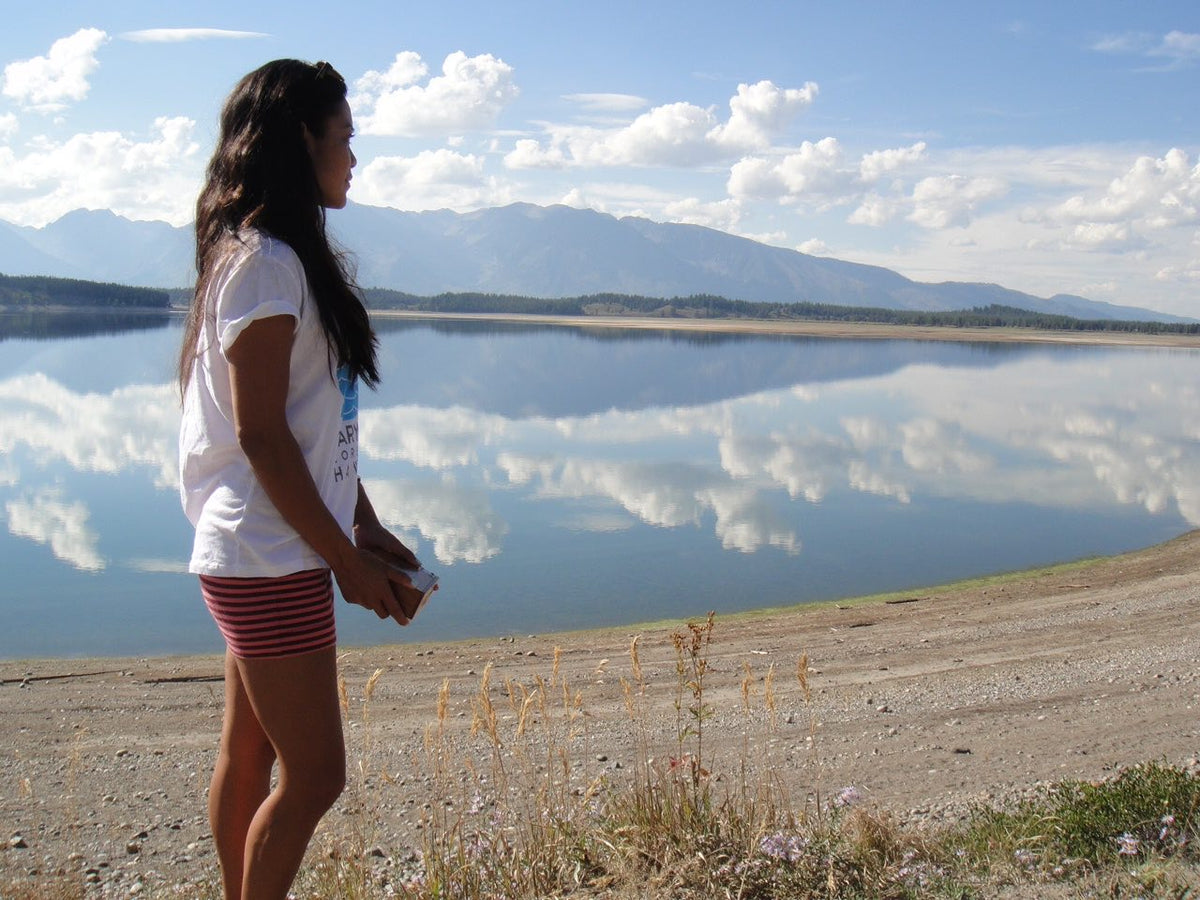 A person with long hair stands by the shore of a calm lake, holding a book. They wear a white t-shirt and red striped shorts. Behind them, mountains and clouds are reflected on the lake's surface, creating a serene and picturesque scene.