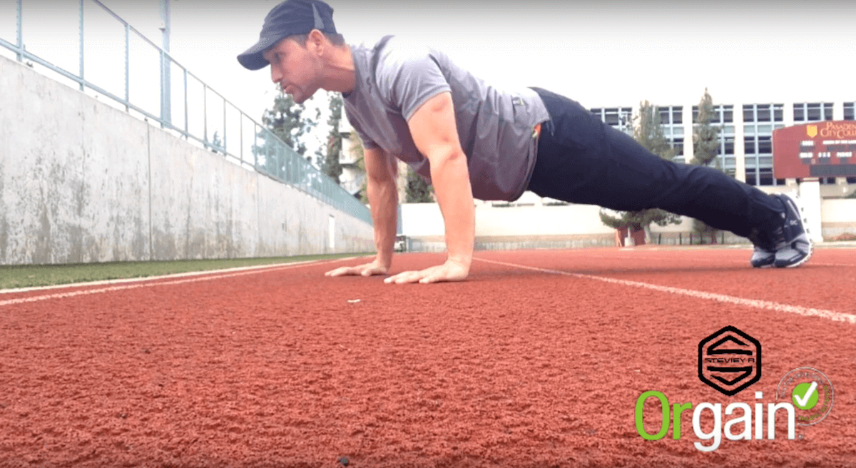 A person in workout attire is performing a plank exercise on a red running track. There is a fence and buildings in the background. The image has a watermark "Orgain" with a checkmark and another blurred logo above it.