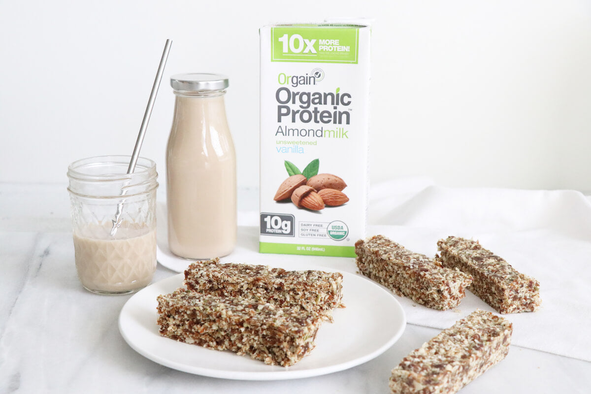 A carton of Orgain Organic Protein Almondmilk stands next to a glass bottle and a small mason jar filled with almond milk. In front, a white plate holds several rectangular nut and seed bars. A metal straw is placed in the mason jar.