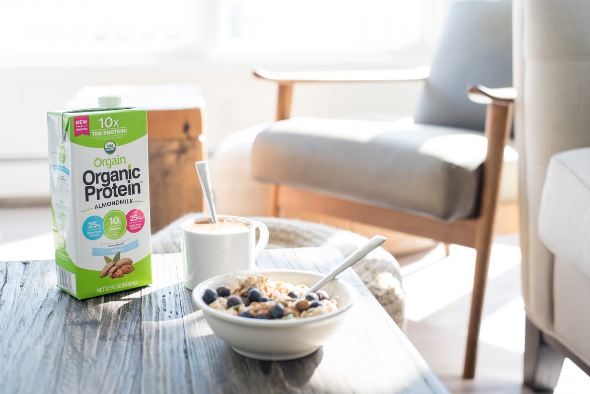 A bowl of blueberries and cereal on the coffee tables, next to a carton of Orgain's Organic Protein.