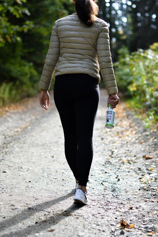 A woman walks down a gravel trail holding an Orgain beverage by the cap with her right hand.