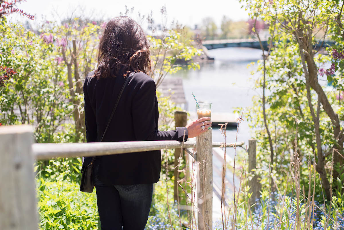 A person with shoulder-length hair, wearing a dark jacket and jeans, stands by a railing holding a cold drink with a straw. They are looking at a scenic view of a river with a distant bridge, surrounded by greenery and colorful flowers.