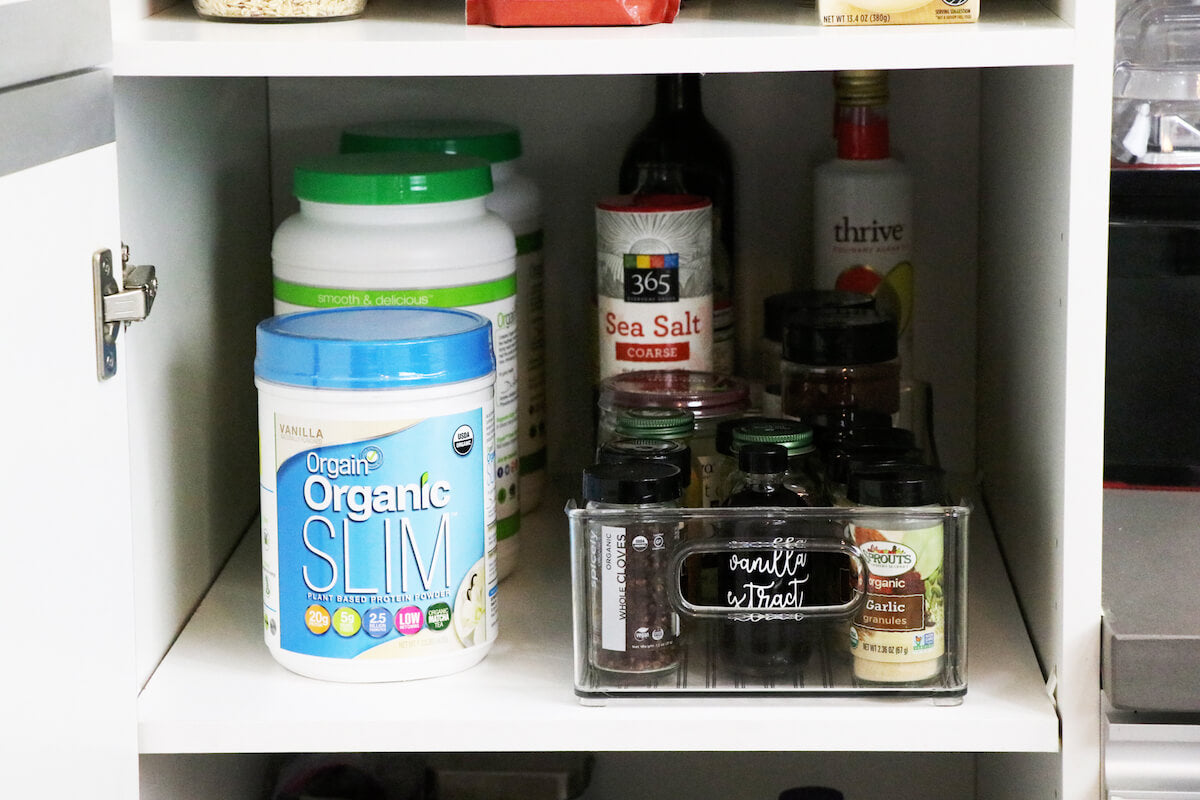 A kitchen cabinet with various items organized inside. On the left shelf, there are containers of protein powder. The middle shelf has sea salt, and various jars and spices are neatly arranged. To the right are more spices in a clear organizer.