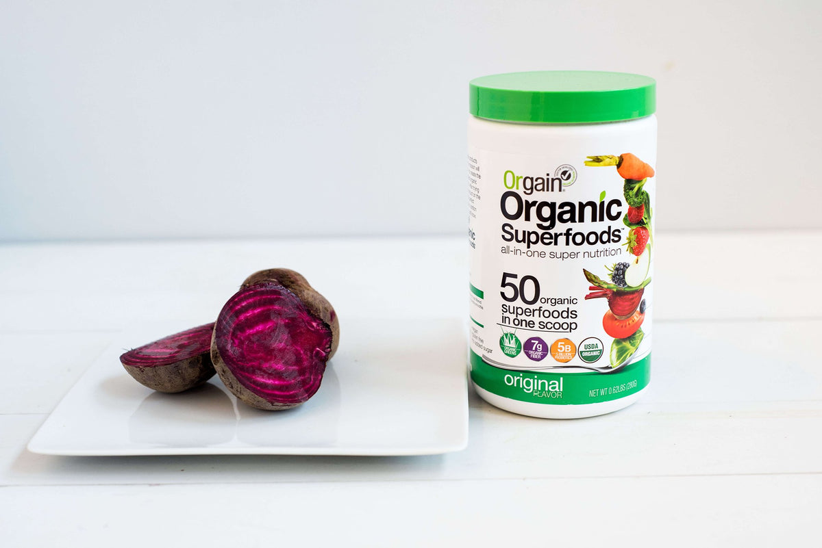 A jar of Orgain Organic Superfoods powder with a green lid is placed on a white surface next to a halved beetroot on a white square plate. The jar is labeled with nutritional information and images of fruits and vegetables.