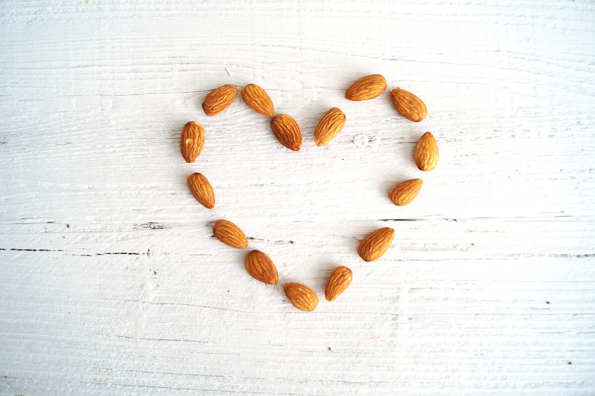 A heart shape made of almonds is arranged on a white wooden surface. The light-colored background contrasts with the brown almonds, which are evenly spaced to form the outline of the heart.