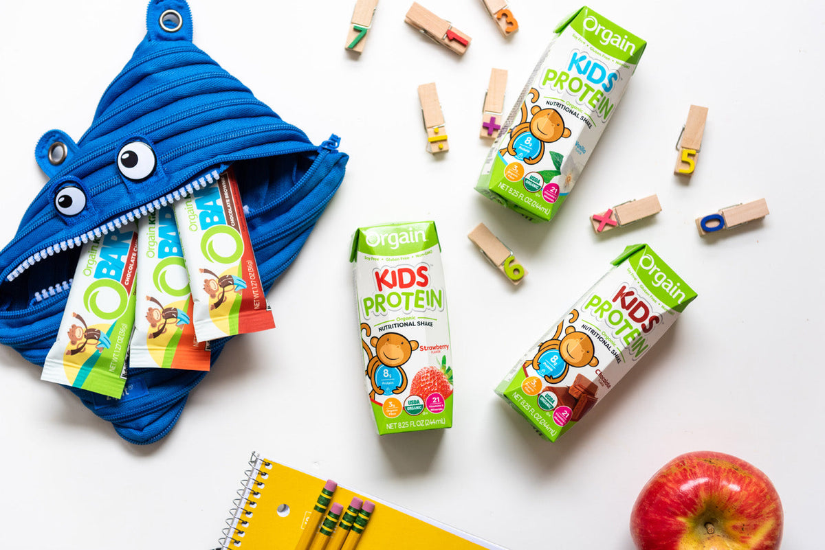 A playful scene with Orgain Kids Protein shakes and bars placed on a white surface. A blue monster-themed pouch holds a few protein bars, while wooden toy pieces, a red apple, and a yellow notebook with colored pencils surround the drinks and pouch.
